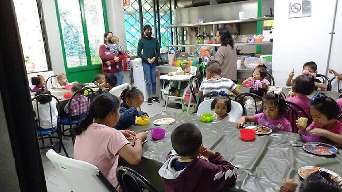 Children sitting at a table eating in a classroom setting with adults supervising.