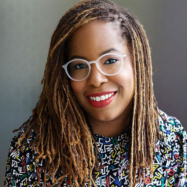 Woman with dreadlocks wearing glasses and a colorful patterned shirt against a gray background