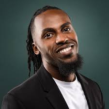 Man with dreadlocks wearing a black jacket over a white shirt against a dark background