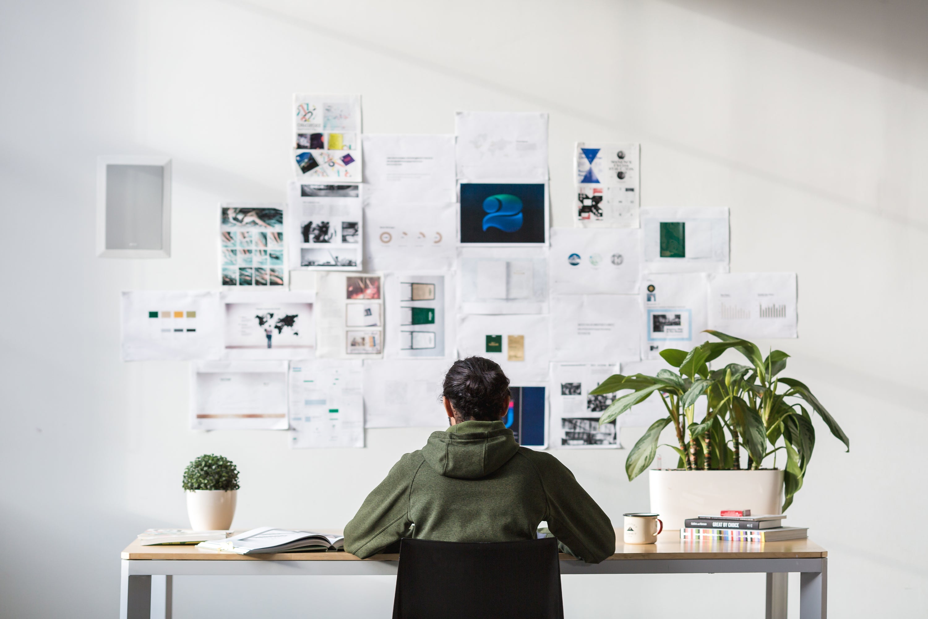 A person in a green hoodie sits at a desk facing a white wall covered with pinned design prints, charts, and mood-board pages. A leafy potted plant, a small shrub, a stack of books, and an enamel mug share the desk in a bright, minimal studio.